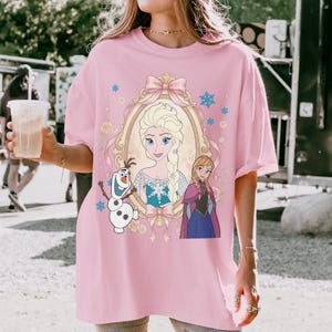 K&ouml;nnte beinhalten: Ein hellrosa T-Shirt mit kurzen &Auml;rmeln und einem Grafikdruck von Elsa, Anna und Olaf aus Die Eisk&ouml;nigin. Elsa ist in einem verzierten goldenen Ovalrahmen mit einer rosa Schleife zentriert, w&auml;hrend Anna und Olaf darunter stehen, umgeben von blauen Schneeflocken.