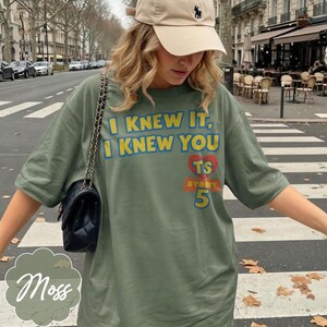 May include: An olive green short-sleeve t-shirt featuring the yellow text I KNEW IT, I KNEW YOU, a red heart with the letters TS, and the word STORY with the number 5 in yellow. A beige baseball cap and a black quilted shoulder bag are also visible.