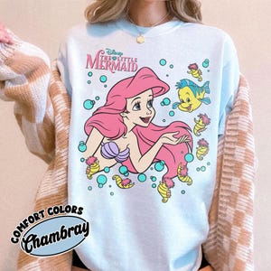 Puede incluir: Una sudadera de cuello redondo azul claro con un gr&aacute;fico de Ariel de La Sirenita con cabello rosa, un top de conchas morado y el pez Flounder. El dise&ntilde;o incluye burbujas de color turquesa y peque&ntilde;os caballitos de mar amarillos. El texto dice Disney The Little Mermaid.