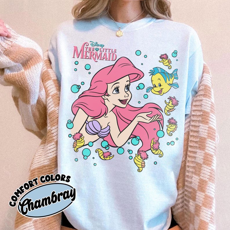 Puede incluir: Una sudadera de cuello redondo azul claro con un gr&aacute;fico de Ariel de La Sirenita con cabello rosa, un top de conchas morado y el pez Flounder. El dise&ntilde;o incluye burbujas de color turquesa y peque&ntilde;os caballitos de mar amarillos. El texto dice Disney The Little Mermaid.