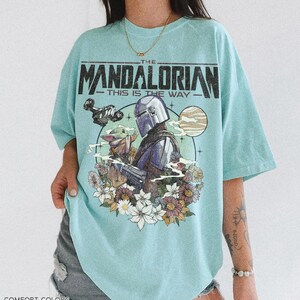 Op de afbeelding: Een lichtblauw T-shirt met een afbeelding van The Mandalorian en Grogu omringd door bloemen. De tekst "THE MANDALORIAN - THIS IS THE WAY" staat bovenaan. Het shirt is van het merk Comfort Colors in de kleur "Charky Mint".