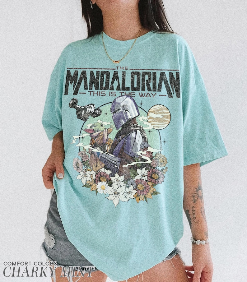 Op de afbeelding: Een lichtblauw T-shirt met een afbeelding van The Mandalorian en Grogu omringd door bloemen. De tekst "THE MANDALORIAN - THIS IS THE WAY" staat bovenaan. Het shirt is van het merk Comfort Colors in de kleur "Charky Mint".