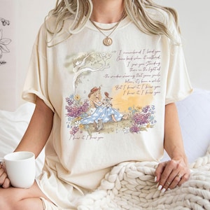 Puede incluir: Una camiseta de manga corta color crema con un gr&aacute;fico de una persona con vestido azul y botas vaqueras sentada en un campo de flores. El texto dice: I remembered I loved you. Come back when it mattered. I saw you standing there in the light of the window wearing that same smile. Man, it's been a while. But I knew it, I knew you. I knew it, I knew you.