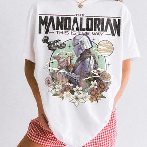 Op de afbeelding: Wit T-shirt met een afbeelding van The Mandalorian, Grogu en een ruimteschip, omringd door bloemen en de tekst "THE MANDALORIAN - THIS IS THE WAY." Het shirt heeft ook de tekst "COMFORT COLORS WHITE" onderaan.