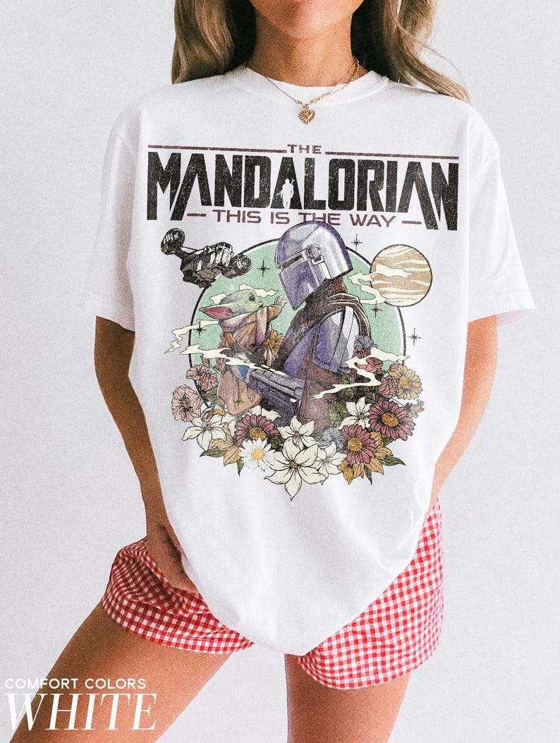 Op de afbeelding: Wit T-shirt met een afbeelding van The Mandalorian, Grogu en een ruimteschip, omringd door bloemen en de tekst "THE MANDALORIAN - THIS IS THE WAY." Het shirt heeft ook de tekst "COMFORT COLORS WHITE" onderaan.