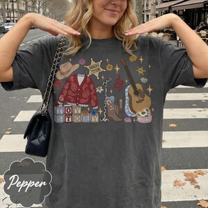 Pode incluir: Uma t-shirt cinzento-carv&atilde;o de manga curta com uma estampa gr&aacute;fica de um chap&eacute;u de cowboy, um casaco de malha vermelho com padr&atilde;o paisley, uma estrela de xerife com o texto Sheriff Taylor, um microfone, uma guitarra e uma bota de cowboy.