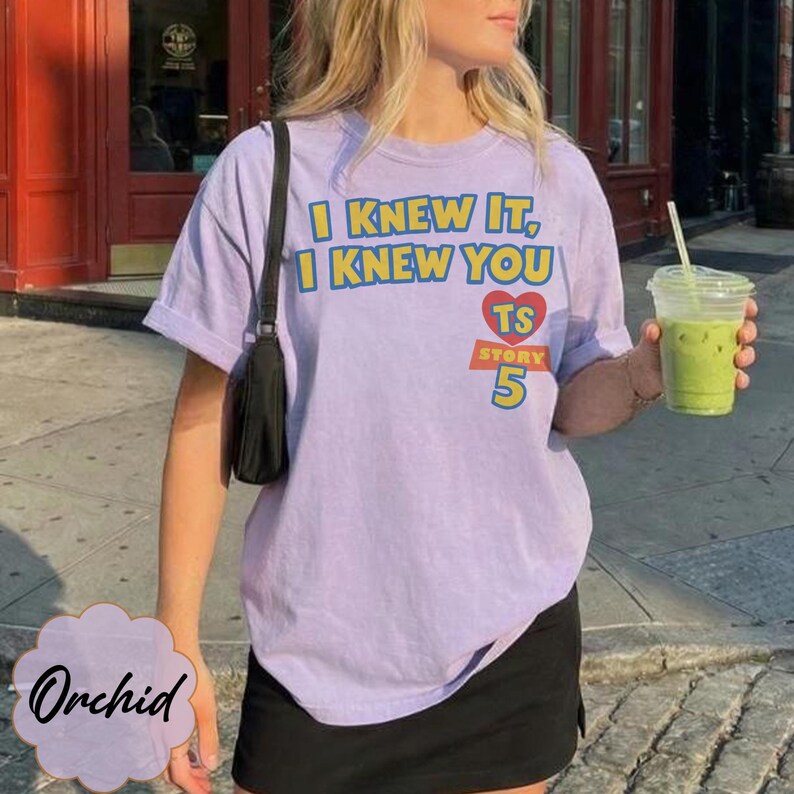 May include: A light purple short-sleeve t-shirt featuring the yellow text I KNEW IT, I KNEW YOU, a red heart with the letters TS, and the text STORY 5 in yellow and orange. The shirt is paired with a black shoulder bag and a black skirt.