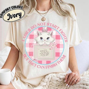 Könnte beinhalten: Ein elfenbeinfarbenes T-Shirt mit einer Grafik eines weißen Kätzchens mit einer rosa Schleife. Das Design enthält einen rosa-weiß karierten Kreis, einen rosa Umschlag und den Text: "LADIES DO NOT START FIGHTS BUT THEY CAN FINISH THEM!" und "always find time for the things that make you feel happy. To be alive."
