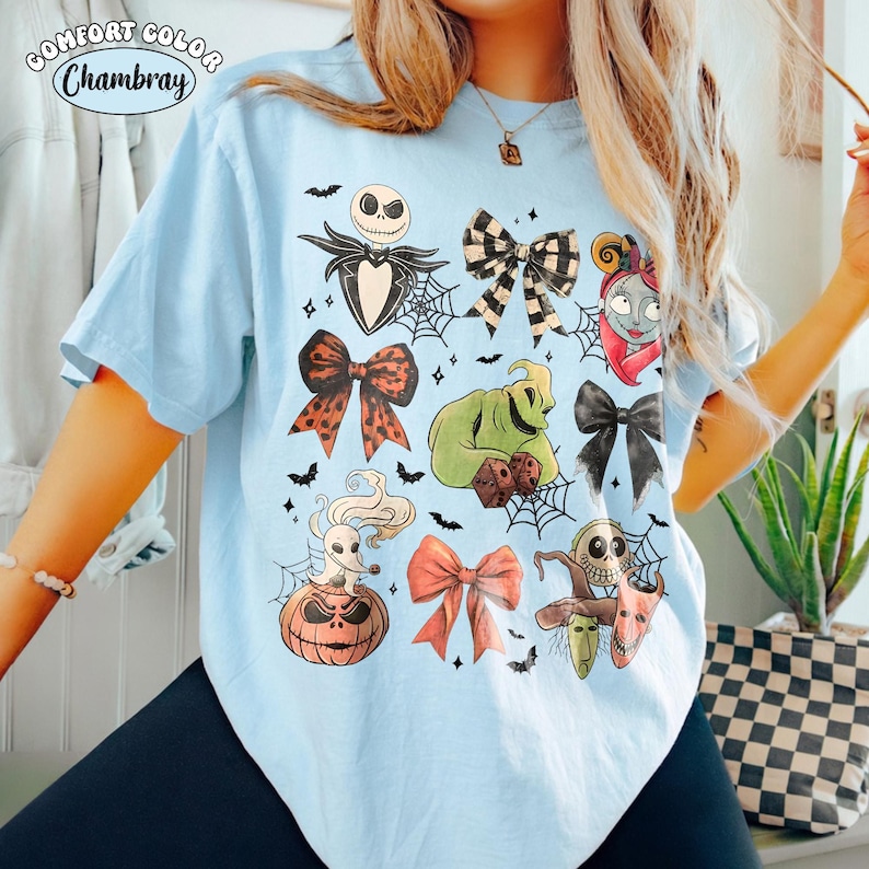 Op de afbeelding: Een lichtblauw t-shirt met een grafische print van personages en strikken uit The Nightmare Before Christmas, waaronder Jack Skellington, Sally, Oogie Boogie, Zero en diverse spinnenwebben, met de tekst Comfort Color Chambray.