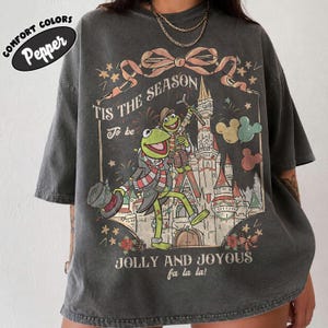 May include: A charcoal gray short-sleeve t-shirt featuring a graphic of Kermit the Frog and Fozzie Bear in front of a castle. The text reads 'TIS THE SEASON To be JOLLY AND JOYOUS fa la la!' with a bow, stars, and Mickey-shaped balloons.