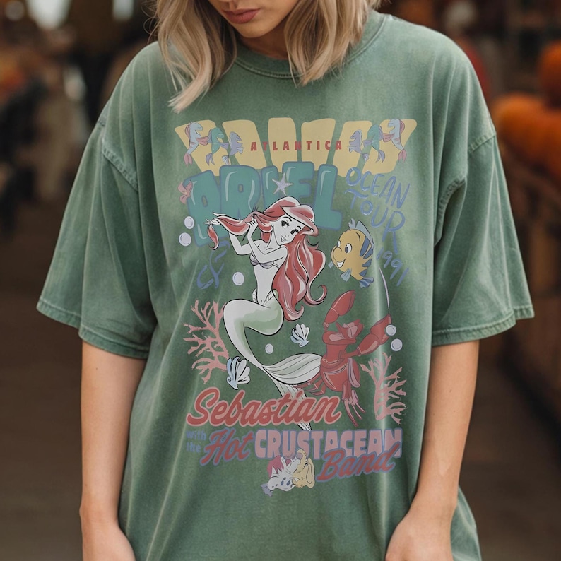 May include: A sage green short-sleeve t-shirt featuring a graphic print of Ariel, Flounder, and Sebastian. The text reads Atlantica Ocean Tour 1991, Sebastian and the Hot Crustacean Band. The design includes coral, bubbles, and shells in muted tones.