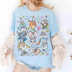 Op de afbeelding: Lichtblauw T-shirt met cartoonfiguren uit Zootopia, elk met een feesthoedje. Het shirt bevat personages als Judy Hopps, Nick Wilde en Flash, met hun namen erboven gedrukt. Het ontwerp is omgeven door sterren en andere feestelijke elementen.