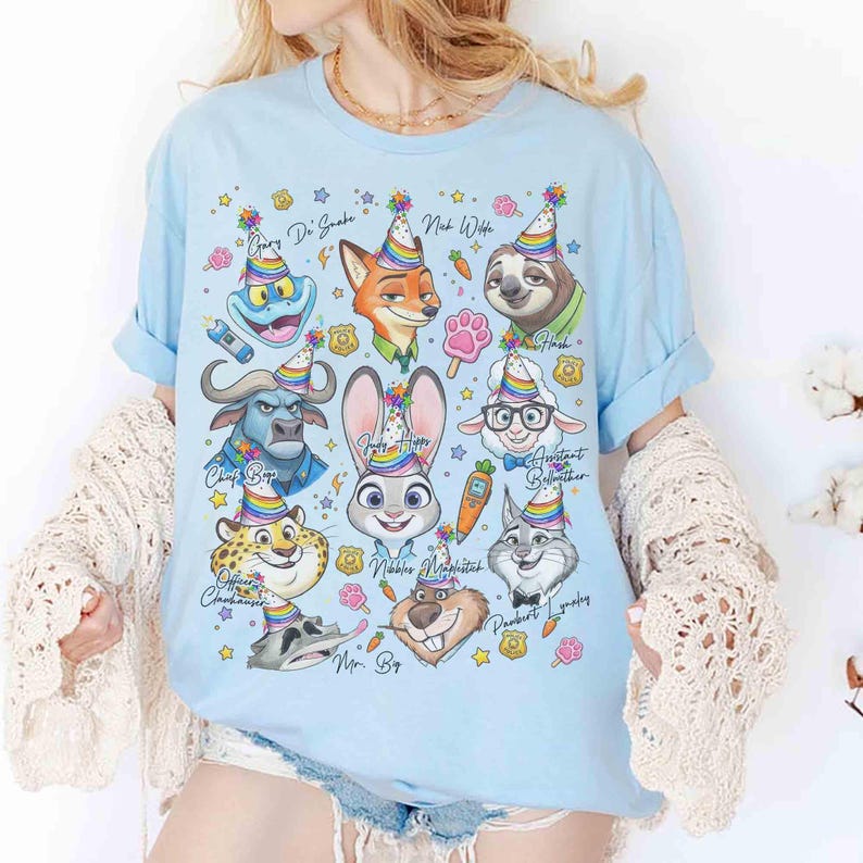 Op de afbeelding: Lichtblauw T-shirt met cartoonfiguren uit Zootopia, elk met een feesthoedje. Het shirt bevat personages als Judy Hopps, Nick Wilde en Flash, met hun namen erboven gedrukt. Het ontwerp is omgeven door sterren en andere feestelijke elementen.