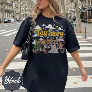 May include: A black short-sleeved t-shirt featuring a graphic print with the text Tay Story in yellow letters. The design includes illustrations of various characters, white clouds, yellow stars, musical notes, and a small rocket on a black background.