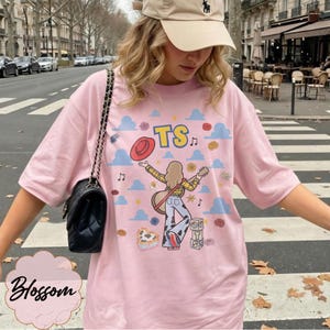 May include: A light pink t-shirt featuring a graphic illustration of a person with long hair, a yellow plaid shirt, and cow-print pants, holding a guitar and a red hat. The design includes blue clouds, musical notes, and blocks with the letters TS.