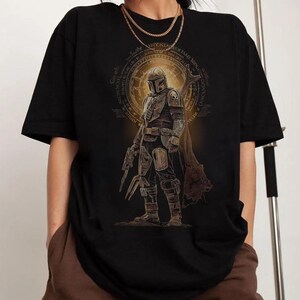 May include: A black short-sleeve t-shirt featuring a graphic print of a armored character in a helmet and cape, holding a weapon. The design includes a circular golden emblem with intricate text patterns surrounding the central figure.