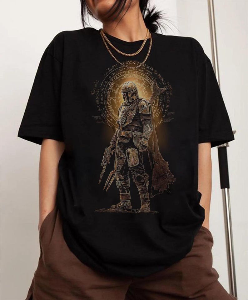 May include: A black short-sleeve t-shirt featuring a graphic print of a armored character in a helmet and cape, holding a weapon. The design includes a circular golden emblem with intricate text patterns surrounding the central figure.