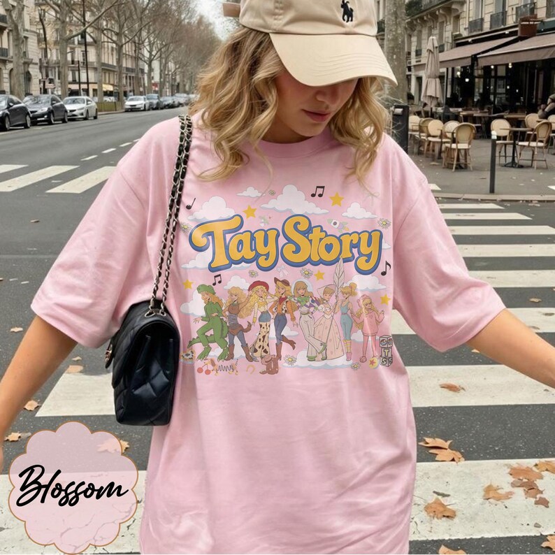 May include: A light pink t-shirt featuring the text Tay Story in yellow with a blue outline. The graphic includes a line of cartoon characters, clouds, stars, and musical notes. A black quilted shoulder bag with a chain strap is worn over the shoulder.