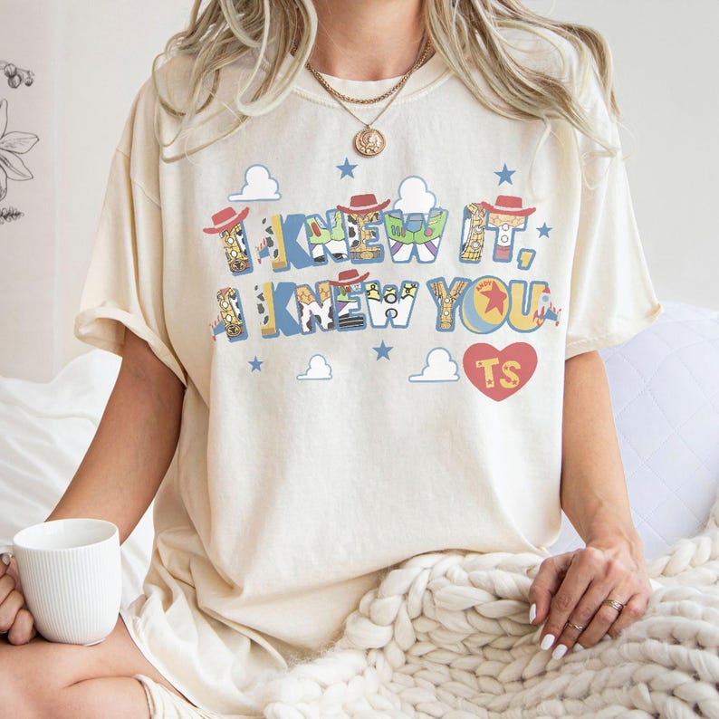 Puede incluir: Una camiseta de manga corta color crema con un estampado gr&aacute;fico que incluye el texto "I KNEW IT, I KNEW YOU" en letras azules y amarillas. El dise&ntilde;o incluye iconos de sombreros de vaquero, nubes, estrellas azules y un coraz&oacute;n rojo con "TS".
