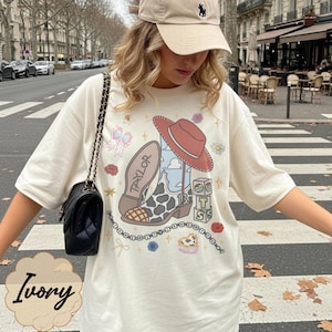 Puede incluir: Una camiseta color marfil con un estampado gr&aacute;fico de una bota vaquera con patr&oacute;n de vaca, un sombrero rojo y el nombre Taylor. El dise&ntilde;o incluye bloques con las letras T, S y un coraz&oacute;n, adem&aacute;s de peque&ntilde;os detalles florales y una cadena de cuentas.