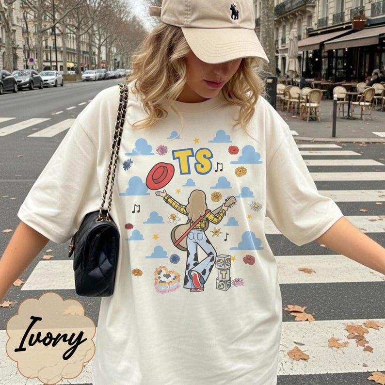 May include: An ivory t-shirt featuring a graphic of a person with a guitar, a red hat, and the text TS. The design includes blue clouds, musical notes, flowers, and cow-print patterns. A black shoulder bag with a chain strap is worn over the shoulder.