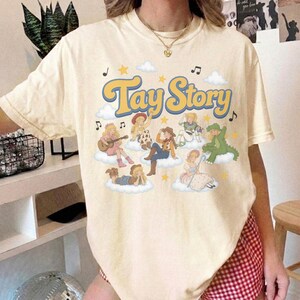 Puede incluir: Una camiseta de manga corta de color crema con el texto Tay Story en letras grandes azules y amarillas. El gr&aacute;fico incluye varios personajes de dibujos animados sentados sobre nubes blancas, rodeados de estrellas amarillas y notas musicales negras.