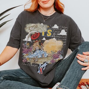 Puede incluir: Una camiseta gris carb&oacute;n con un estampado gr&aacute;fico de una persona con vestido azul y sombrero de vaquero sentada en un campo de flores. El dise&ntilde;o incluye el texto "I knew it, I knew you" y "I saw you there" con un sombrero rojo y nubes.