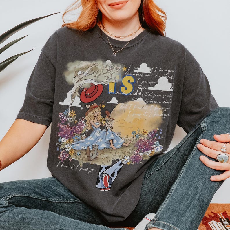 Puede incluir: Una camiseta gris carb&oacute;n con un estampado gr&aacute;fico de una persona con vestido azul y sombrero de vaquero sentada en un campo de flores. El dise&ntilde;o incluye el texto "I knew it, I knew you" y "I saw you there" con un sombrero rojo y nubes.