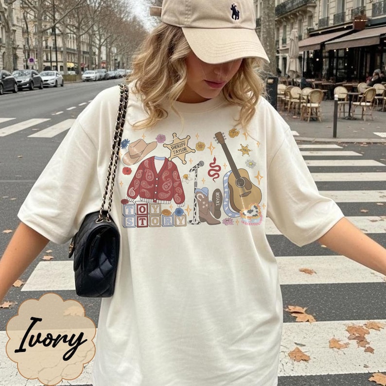 Pode incluir: Uma t-shirt cor de marfim com uma estampa gr&aacute;fica de um chap&eacute;u de cowboy, um cardig&atilde; vermelho com padr&atilde;o paisley, uma estrela de xerife com a inscri&ccedil;&atilde;o 'Sheriff Taylor', um microfone, uma bota de cowboy, uma guitarra e blocos 'Toy Story'.