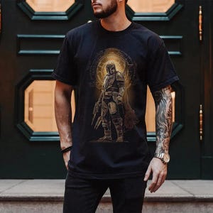 May include: A black short-sleeved t-shirt featuring a graphic print of a armored character in a helmet standing in front of a glowing golden circular emblem. The design includes intricate line work and a weathered, metallic aesthetic.