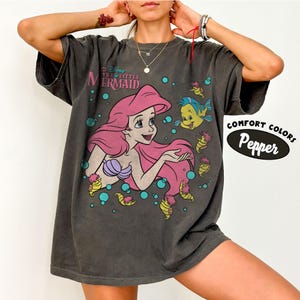 Puede incluir: Una camiseta de manga corta gris carb&oacute;n con un gr&aacute;fico de Ariel de La Sirenita con cabello rosa y un top de conchas morado, acompa&ntilde;ada por Flounder y caballitos de mar peque&ntilde;os entre burbujas azules. El texto dice Disney The Little Mermaid.