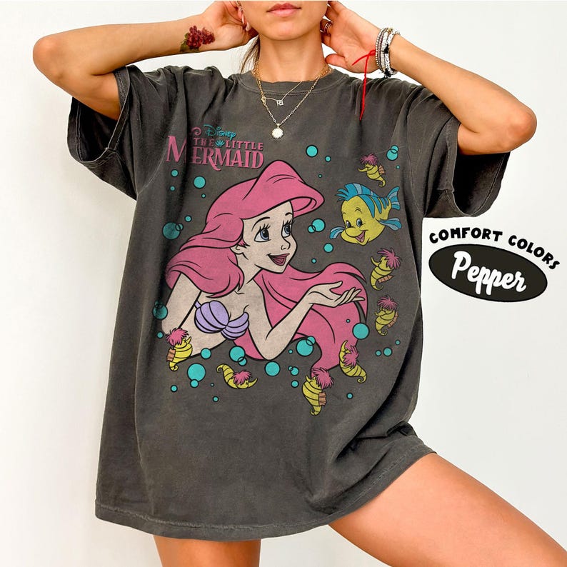 Puede incluir: Una camiseta de manga corta gris carb&oacute;n con un gr&aacute;fico de Ariel de La Sirenita con cabello rosa y un top de conchas morado, acompa&ntilde;ada por Flounder y caballitos de mar peque&ntilde;os entre burbujas azules. El texto dice Disney The Little Mermaid.