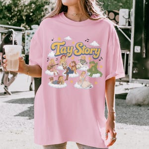 Puede incluir: Una camiseta rosa claro con el texto "Tay Story" en una fuente retro amarilla y azul. El gr&aacute;fico incluye varias figuras ilustradas con diversos atuendos, cada una sentada o de pie sobre nubes blancas, rodeadas de notas musicales.