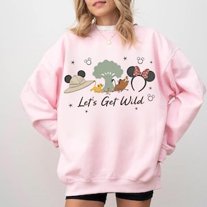Könnte beinhalten: Hellrosa Sweatshirt mit einem Aufdruck, der einen Safarihut, Simba, Timon, Pumbaa und Minnie-Maus-Ohren zeigt. Der Text "Let's Get Wild" ist darunter gedruckt, mit Sternen und Mickey-Mouse-Kopf-Akzenten.