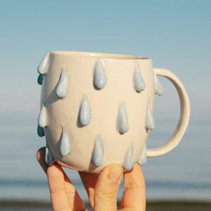 Könnte beinhalten: Ein Set aus drei Keramikbechern mit gesprenkelter, matter Oberfläche. Jeder Becher hat eine andere Farbe: hellblau, cremefarben und grau. Die Becher haben ein schlichtes, modernes Design und einen bequemen Griff.