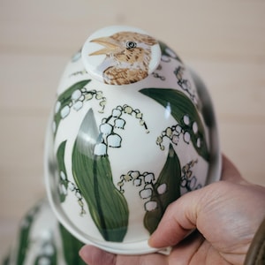 Puede incluir: Cuenco de cerámica blanco con tapa y pomo de pájaro. El cuenco está decorado con hojas verdes y flores blancas de lirios de los valles. La tapa tiene una ilustración detallada de la cabeza de un pájaro.