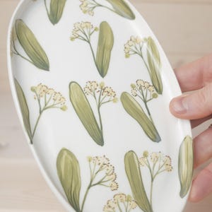 Puede incluir: Plato ovalado de cerámica blanca con un diseño botánico pintado a mano. El diseño presenta hojas verdes y pequeñas flores amarillas. El plato está sostenido por una mano.