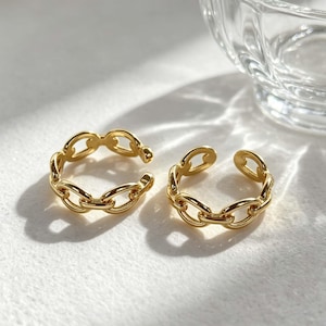 May include: Two gold-colored chain link rings. The rings are open-ended, allowing for adjustable sizing. The rings are displayed on a white surface, with a glass object in the background.