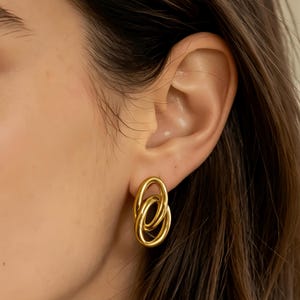 May include: Gold-colored oval stud earrings. The earrings feature a linked design, with two interlocking oval shapes. The earrings are shiny and reflect light. The earrings are a jewelry item.