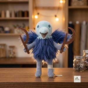 May include: A plush bird toy with a light blue head and a dark blue shaggy body. The bird has a light brown beak and holds two brown, crescent-shaped felt tools in its wings. A small white bandage is wrapped around one of its light blue legs.