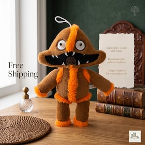 May include: A handmade brown plush toy with orange accents, featuring a unique monster-like design. The toy has large white eyes, a black mouth with teeth, and orange trim. The text "Each item is woven with a story" is visible.