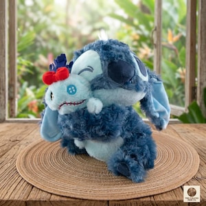Op de afbeelding: Een pluche speelgoed met een blauw, buitenaards personage dat een kleinere versie van zichzelf omhelst. Het grotere pluche heeft grote oren en een vrolijke uitdrukking. Het kleinere pluche heeft een rode strik en een blauwe knoop.