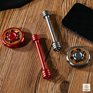 Könnte beinhalten: Eine Sammlung von Metall-Fidget-Spielzeugen in Rot, Silber und Gold. Das Bild zeigt zwei Spinner und zwei stabförmige Fidget-Spielzeuge. Die Spinner haben ein kreisförmiges Design mit mehreren Löchern. Die stabförmigen Spielzeuge sind zylindrisch.
