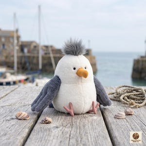 May include: A plush seagull toy with a white body, gray wings, and a gray tuft of feathers on its head. It has a yellow beak and pink feet. The toy is sitting on a wooden surface with seashells.