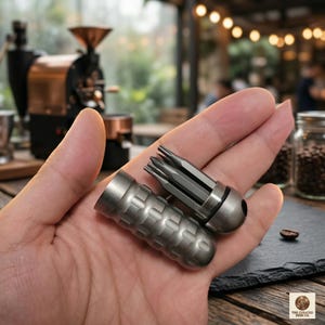 Pode incluir: Um pequeno conjunto de ferramentas cinza metálico com várias pontas, segurado em uma mão. O conjunto inclui uma caixa cilíndrica com um padrão texturizado e um porta-pontas. O fundo apresenta um moedor de café e grãos de café.