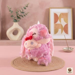 Op de afbeelding: Een roze pluche speelgoed met een rode strik en een kleiner personage dat het omhelst. Het speelgoed heeft lange oren en een lachend gezicht. Het staat op een geweven mat, met een vaas en ingelijste kunst op de achtergrond.