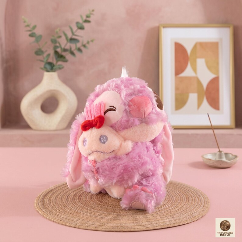 Op de afbeelding: Een roze pluche speelgoed met een rode strik en een kleiner personage dat het omhelst. Het speelgoed heeft lange oren en een lachend gezicht. Het staat op een geweven mat, met een vaas en ingelijste kunst op de achtergrond.