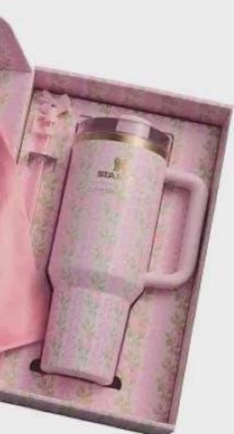 Puede incluir: Un vaso Stanley rosa con estampado floral y asa, presentado en una caja de regalo a juego. El vaso tiene un borde dorado y el logotipo de Stanley. La caja de regalo tambi&eacute;n es rosa y floral.
