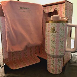 Puede incluir: Un termo Stanley con estampado floral y asa, presentado en una caja a juego. El termo presenta un dise&ntilde;o floral rosa y blanco, detalles dorados y el logotipo "Stanley Loveshackfancy". Tambi&eacute;n se incluye una bolsa de tela rosa.