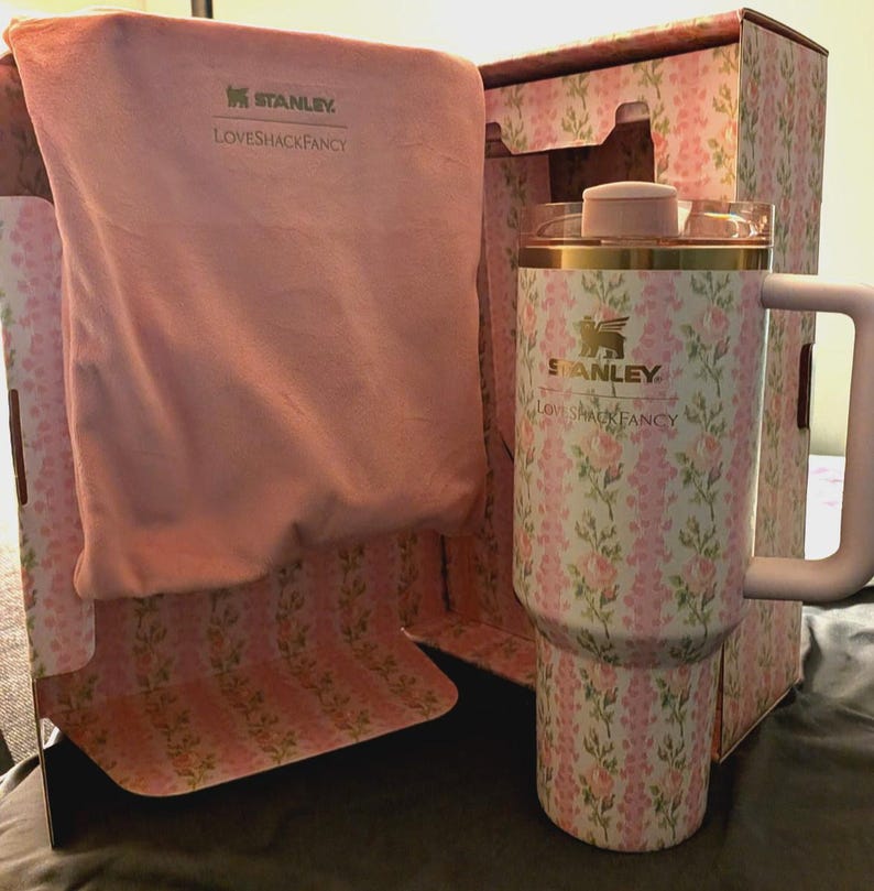 Puede incluir: Un termo Stanley con estampado floral y asa, presentado en una caja a juego. El termo presenta un dise&ntilde;o floral rosa y blanco, detalles dorados y el logotipo "Stanley Loveshackfancy". Tambi&eacute;n se incluye una bolsa de tela rosa.